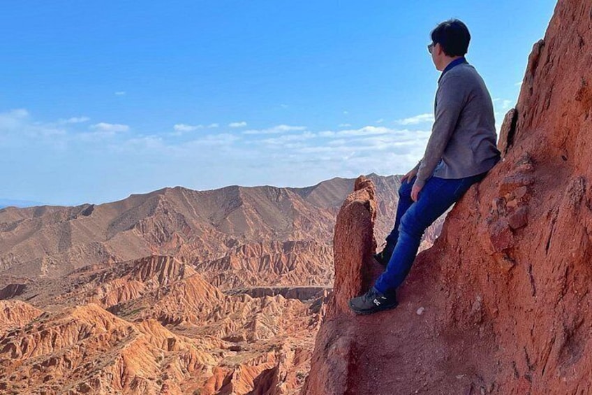 A person sitting on a red rock ledge overlooking a rugged mountainous landscape. 2