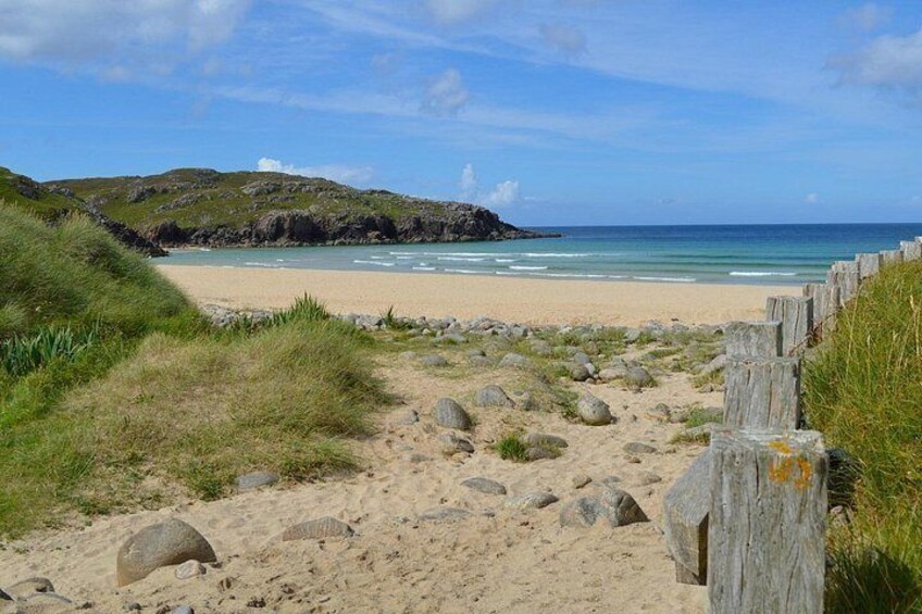 A sandy beach with a wooden fence, grassy dunes, and a rocky outcrop in the distance. 2