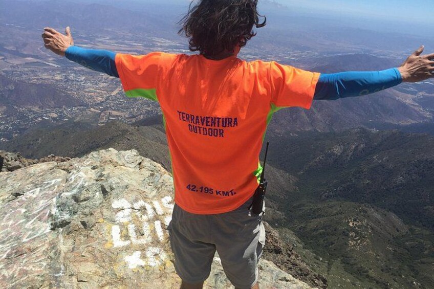 Een persoon staat op een bergtop met uitgestrekte armen, gekleed in een oranje shirt met de tekst 'TERRAVENTURA OUTDOOR' en het cijfer '42.195 KM' op de rug. 1