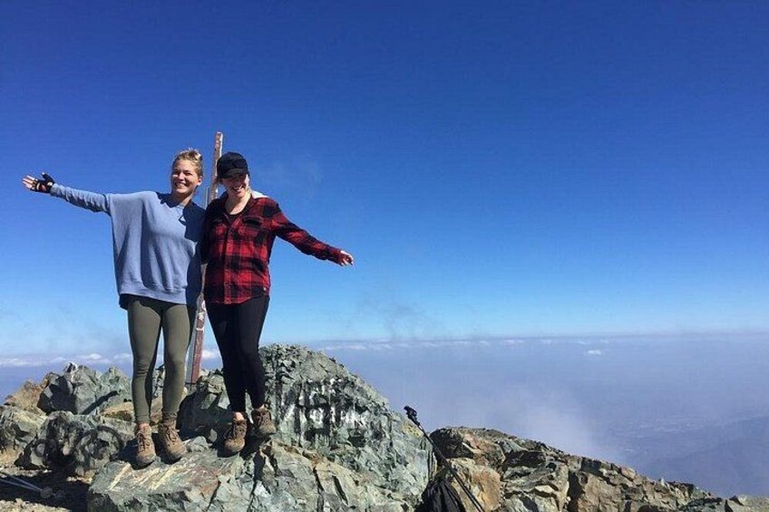 Twee personen staan op een rotsachtige bergtop, met een heldere lucht op de achtergrond. 3