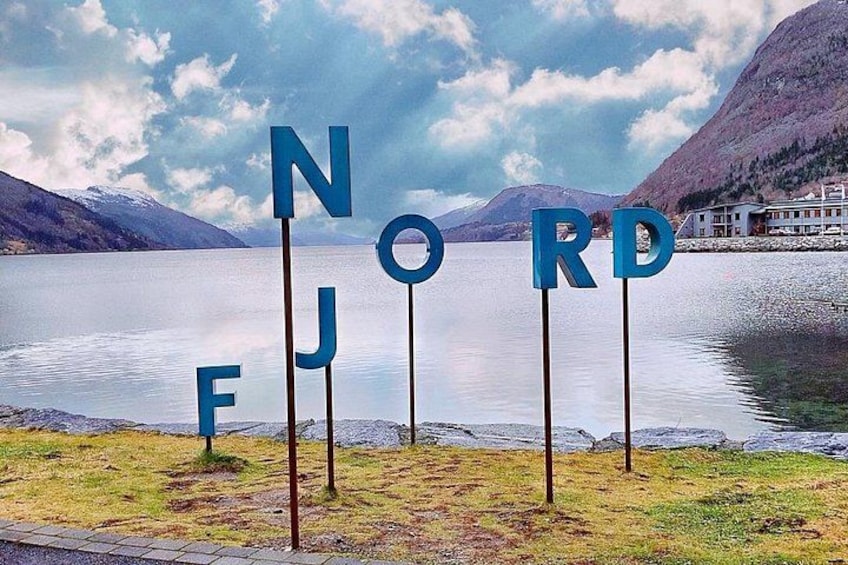 A lake with mountains in the background and the word 'NORD' spelled out in large letters on the shore. 1