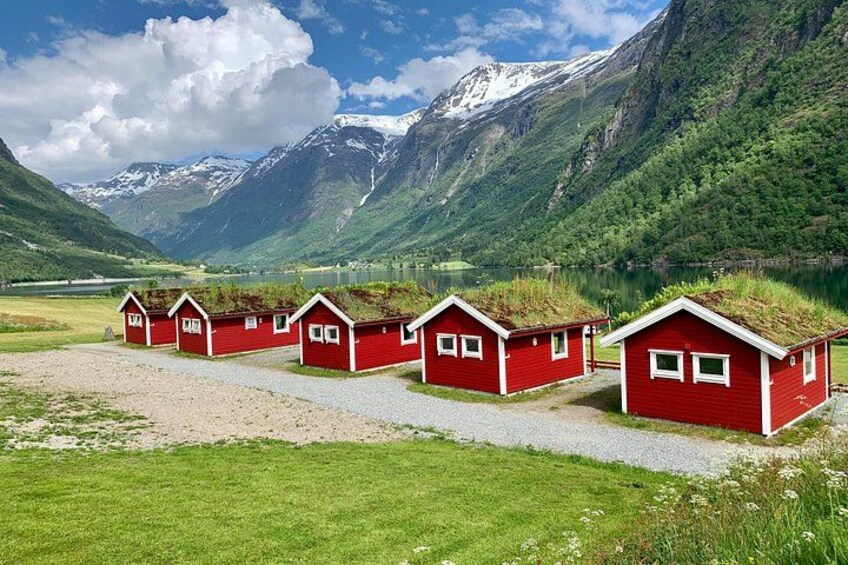 A row of red cabins with grass roofs, a gravel path, and a mountainous landscape with a lake and snow-capped peaks in the background. 5