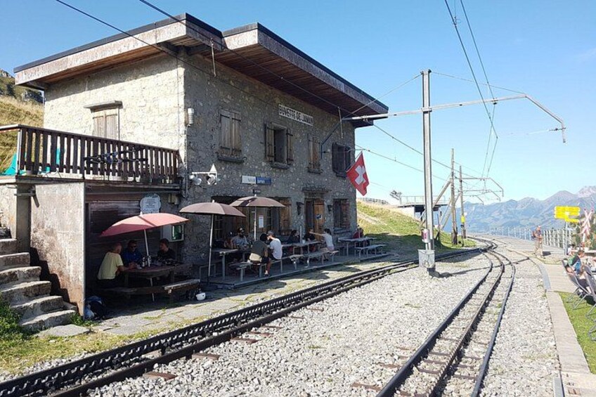 A train station with a Swiss flag, outdoor seating, and a mountain backdrop. 2