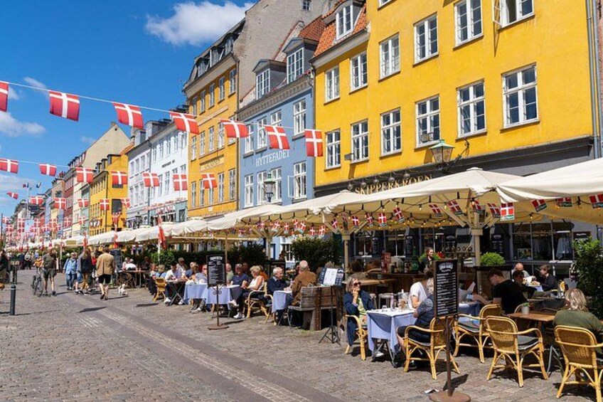 A cobblestone street lined with colorful buildings, outdoor seating areas, and Danish flags. 1