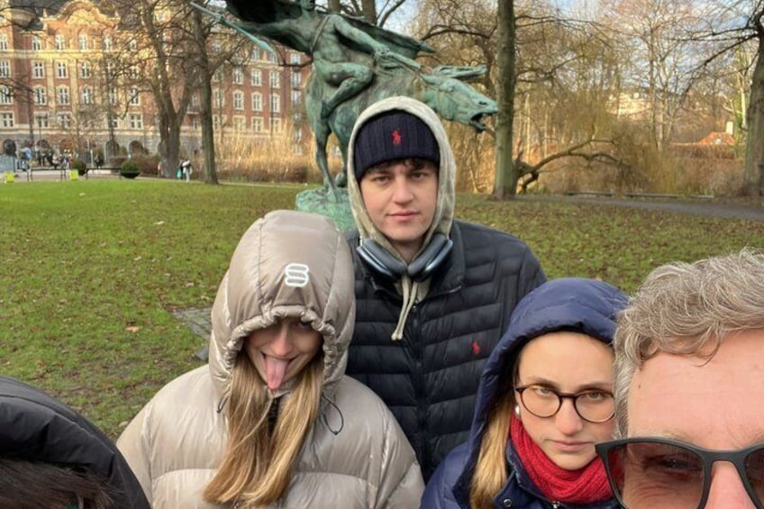 En grupp människor i en park, med en staty och byggnader i bakgrunden. 3