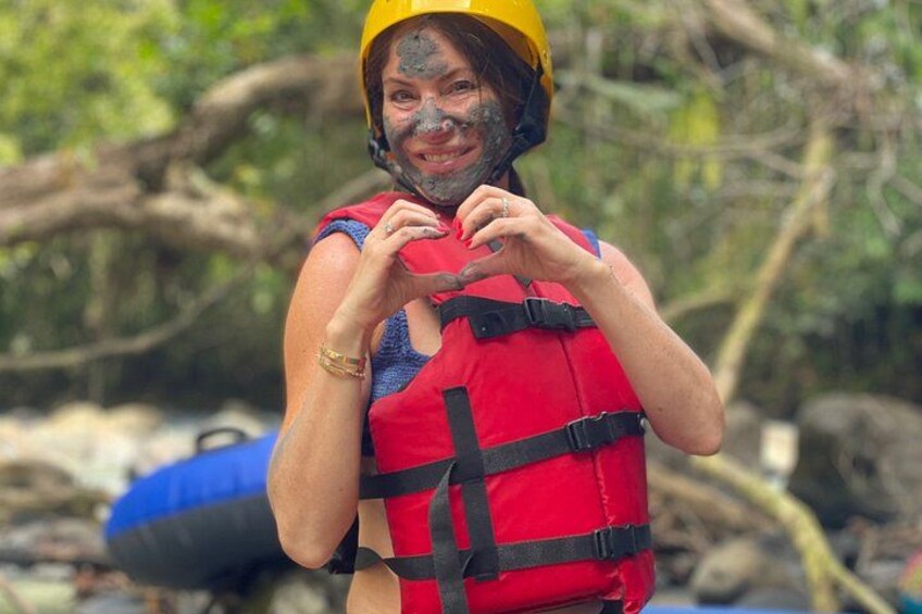 A person wearing a yellow helmet and a red life jacket, standing outdoors with trees and a blue object in the background. 4