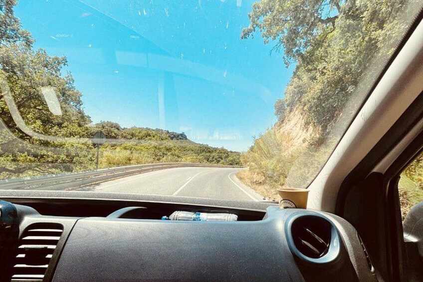 Le tableau de bord d’une voiture et son pare-brise, avec une route qui serpente devant, le tout entouré de verdure et sous un ciel dégagé. 2