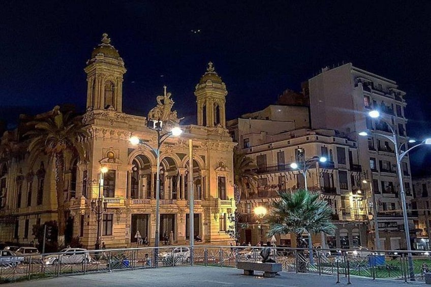 A historic building with ornate architecture and two towers, surrounded by modern buildings and street lamps. 1