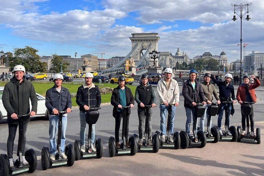 En grupp människor på segways i en stadsmiljö, med en bro och byggnader i bakgrunden. 1