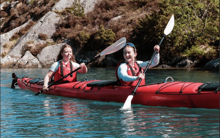 Two people kayaking in a red kayak on a calm river with rocks and greenery in the background. 2