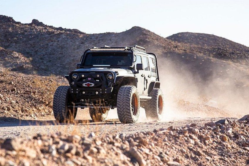 A rugged off-road vehicle driving on a dirt road with a rocky terrain and hills in the background. 3