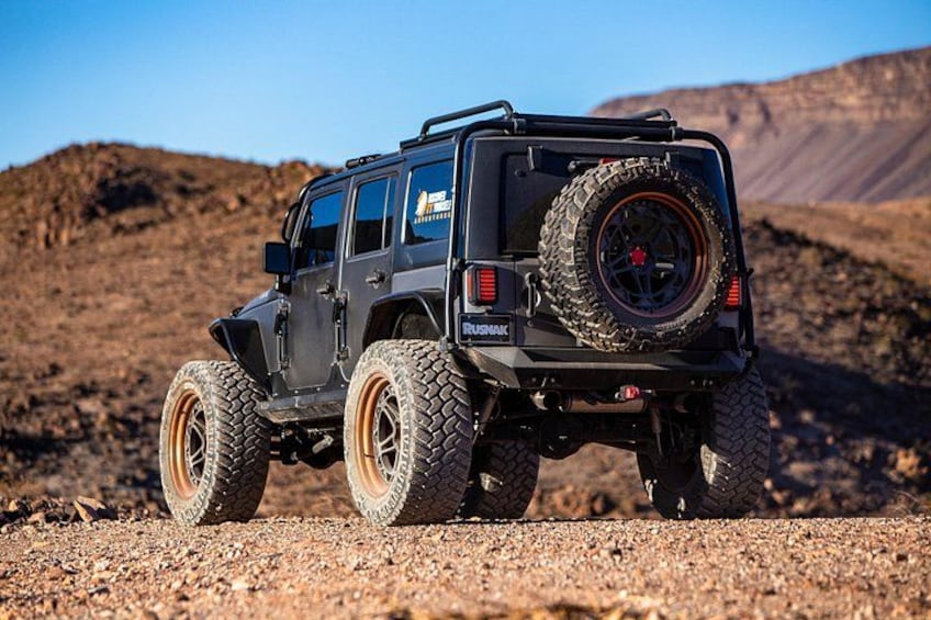 A black off-road vehicle with large tires and a spare tire mounted on the back, parked on a rocky terrain with a desert landscape in the background. 2