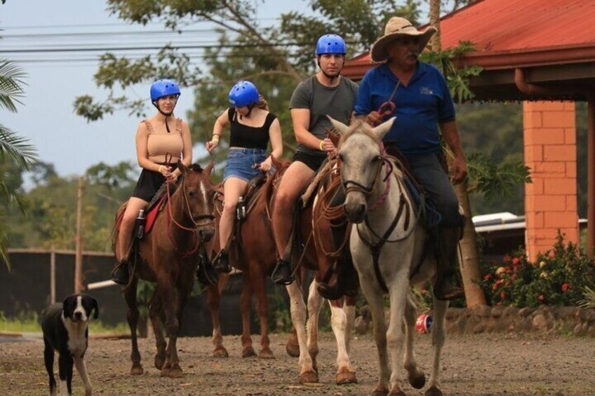 Trois personnes montent à cheval sur un chemin de terre, avec un bâtiment en briques en arrière-plan. 3