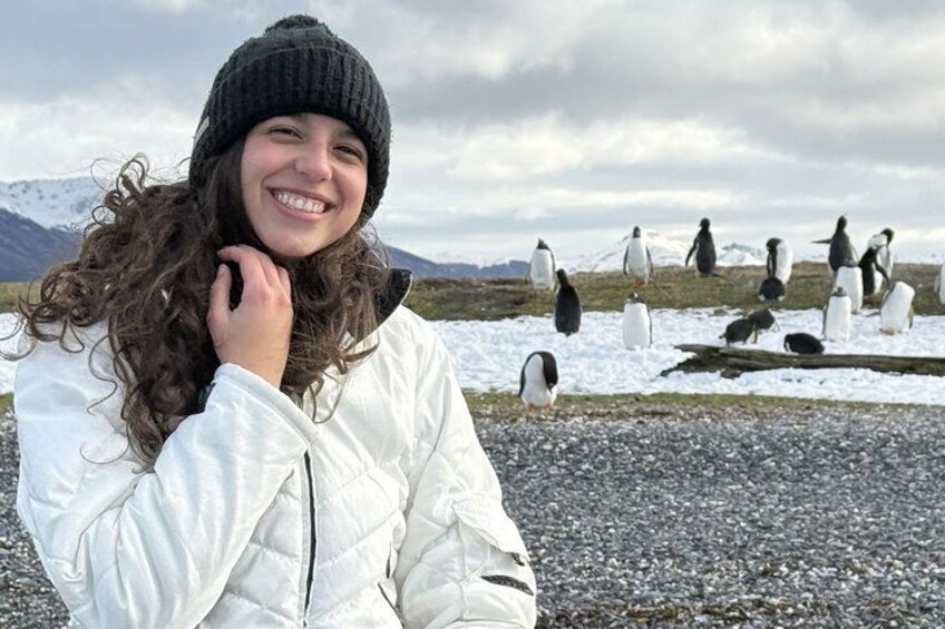 Una persona con chaqueta blanca y gorro negro, de pie en terreno pedregoso con pingüinos al fondo. 1
