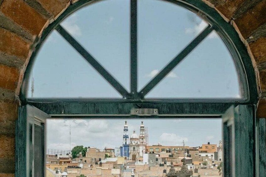 Blick durch ein Fenster mit Ziegelbogen auf eine Stadtansicht mit Gebäuden und einem markanten Turm. 2