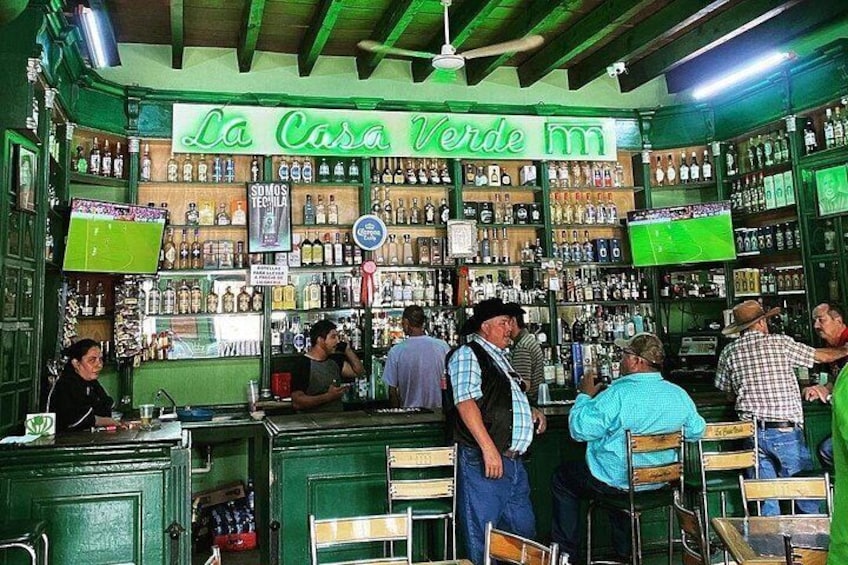 Un bar con decoración interior verde y un letrero que dice 'La Casa Verde'. Hay estantes con botellas y una pantalla de televisión. 2