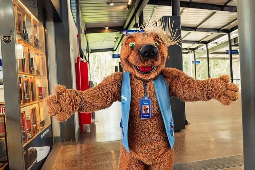 A mascot with a blue vest and ID badge in a covered indoor area with shelves and a red fire extinguisher. 2