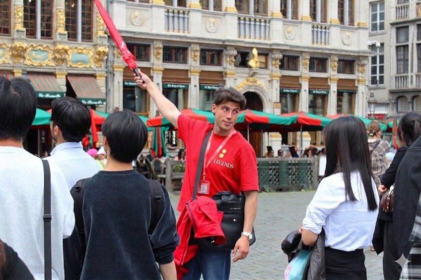 Een persoon in een rood shirt houdt een rood voorwerp vast, staand in een druk open gebied met gebouwen op de achtergrond. 1