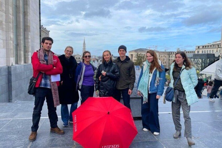 Un groupe de personnes se tient sur une surface pavée, et un parapluie rouge est visible au premier plan. 3