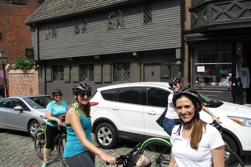 A group of people with bicycles, a white car, and a building with a brick facade. 2