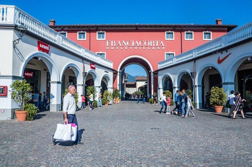 A cobblestone street with arches, a red building labeled 'FRANCIACORTA OUTLET VILLAGE,' and a person carrying a shopping bag. 1