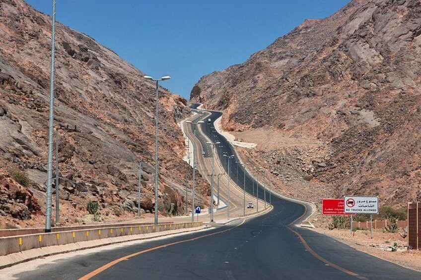 A winding mountain road with guardrails, a clear sky, and rocky terrain. 1