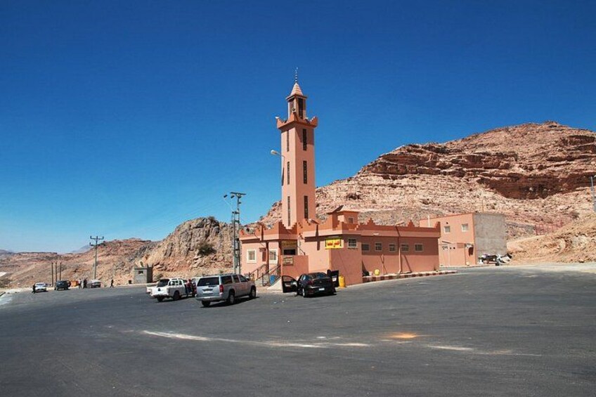 A desert landscape with a prominent building featuring a tall tower, surrounded by a paved area with parked vehicles. 2