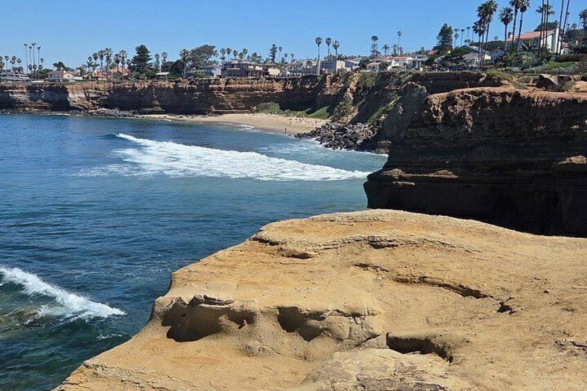 A coastal landscape with a rocky shoreline, waves, and a distant residential area with palm trees. 2