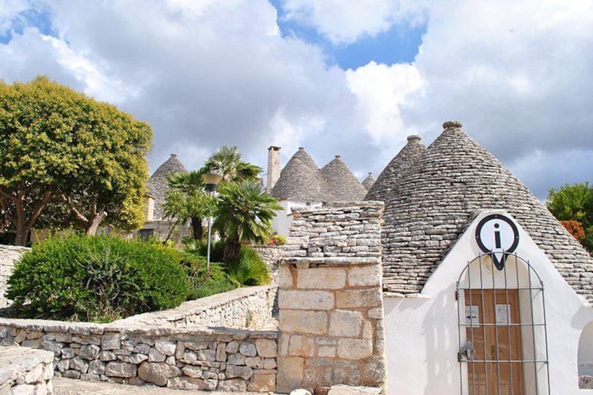 Un edificio in pietra con un tetto a punta, un cancello con il simbolo di informazione e un muro di pietra con vegetazione. 1