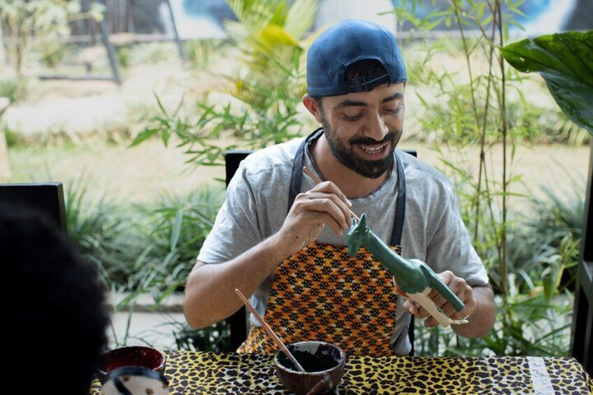 Una persona con gorra y delantal sosteniendo un objeto verde, con una mesa y plantas al fondo. 3