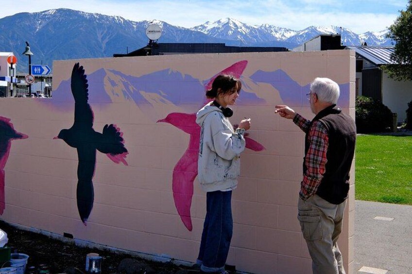 Deux personnes se tiennent devant un mur orné d’oiseaux peints, avec des montagnes en arrière-plan. 5