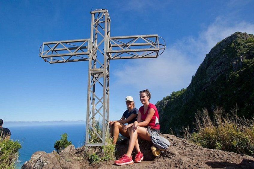 Two people sitting on a rocky outcrop with a large metal cross structure behind them, overlooking a coastal landscape. 3