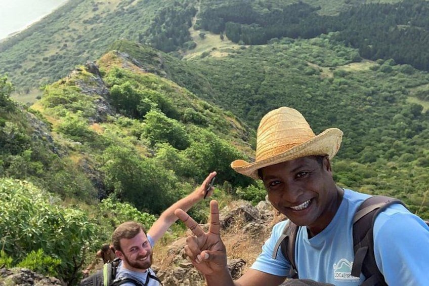 A person wearing a straw hat and a blue shirt, with a backpack, standing on a mountain with greenery and rocky terrain. 5