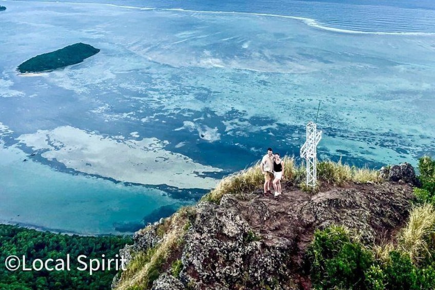 A couple on a rocky outcrop with a cross structure, overlooking a turquoise sea with scattered islands. 1