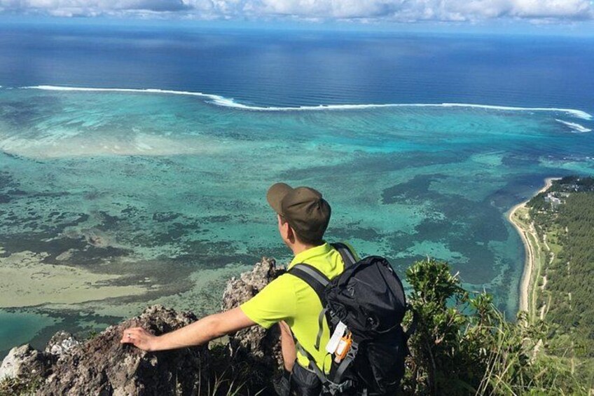 A person in a yellow shirt and brown hat sitting on a rocky outcrop overlooking a turquoise sea with clear waters and a sandy beach. 2