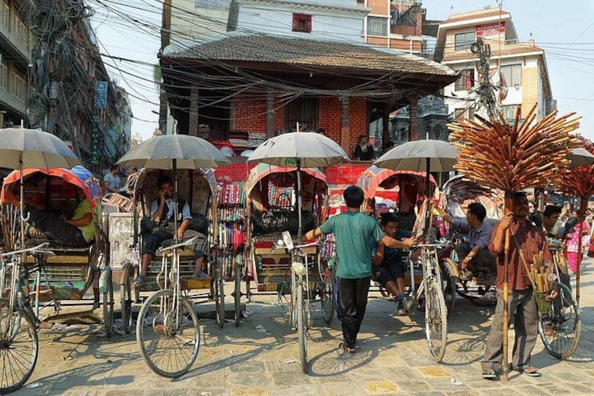 Escena callejera con riquishás y bicicletas, gente sentada y de pie, y un edificio de techo rojo al fondo. 4