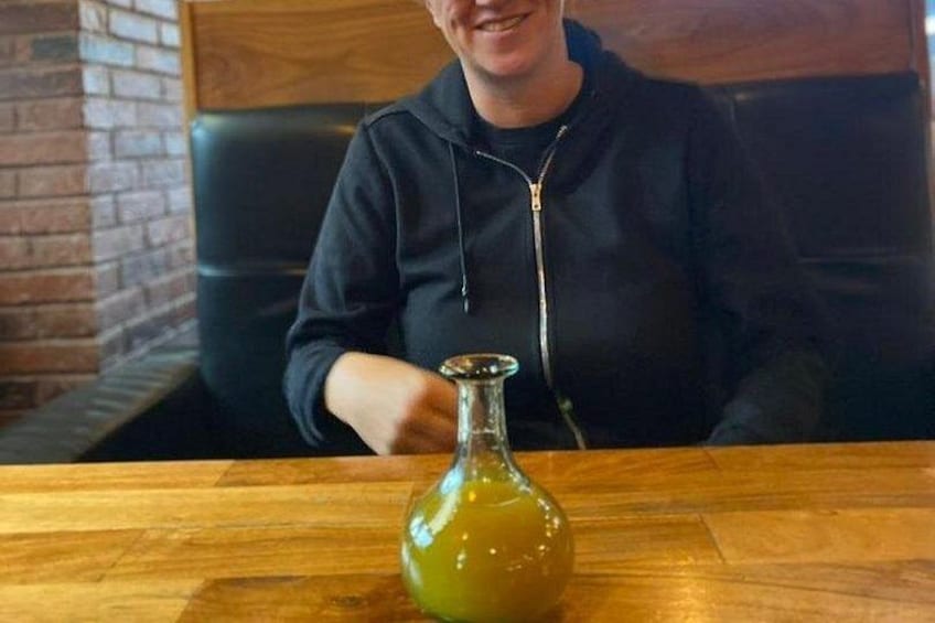 Een persoon zit aan een houten tafel met een groene fles voor zich. 3