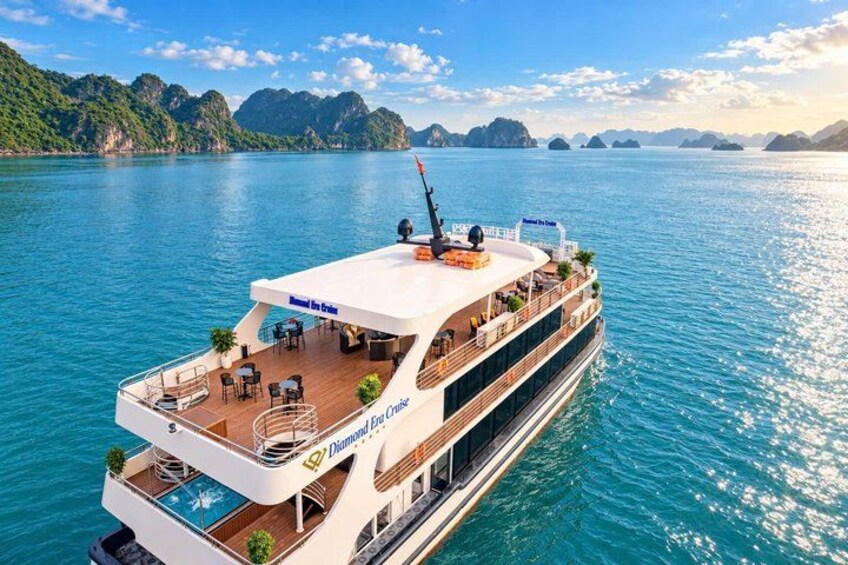 A large cruise ship with a swimming pool and outdoor seating on the upper deck, sailing in a calm sea with mountainous coastline in the background. 1