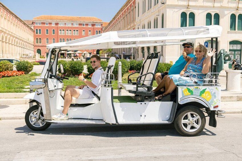 A golf cart with passengers, parked on a street with buildings and greenery in the background. 2