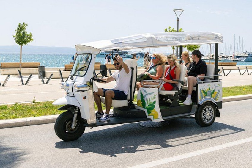 A white golf cart with passengers, parked on a road with a waterfront and boats in the background. 3