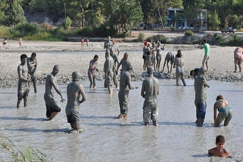 Personas vadeando en aguas poco profundas, algunas con barro en el cuerpo, en un entorno natural con árboles y un edificio al fondo. 4