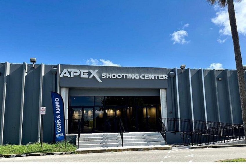A building with a sign that reads 'APEX SHOOTING CENTER' and a blue flag with 'GUNS & AMMO' written on it. 2
