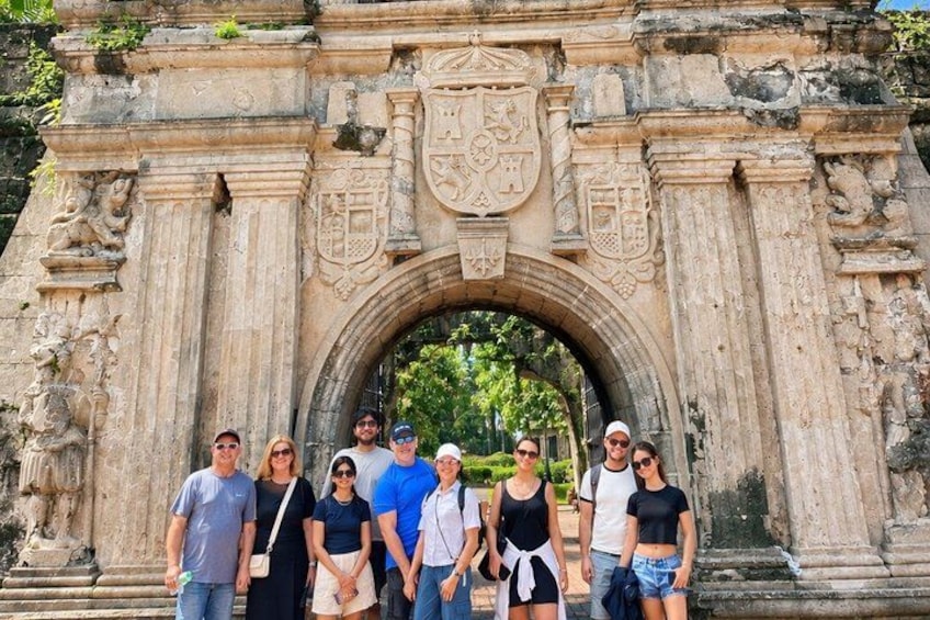 Un grupo de personas posando frente a un arco de piedra histórico con intrincados adornos y un escudo de armas. 1