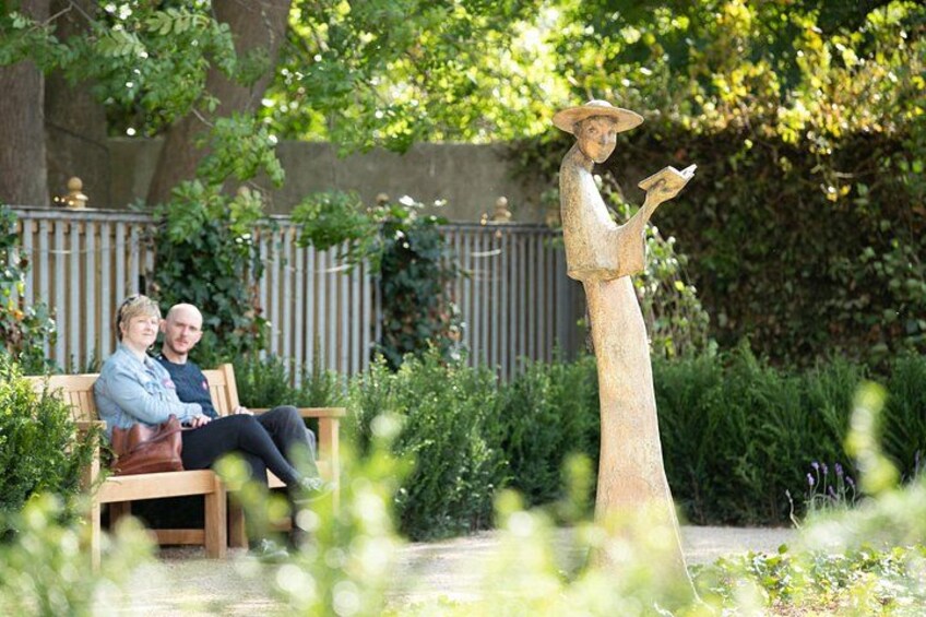 A couple sitting on a bench, a statue of a person in a hat, and a garden with greenery and a fence in the background. 3