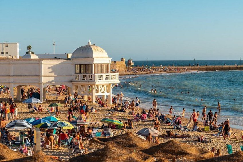 Una playa concurrida, gente tomando el sol y nadando, un edificio blanco con cúpula, y un cielo azul despejado. 5