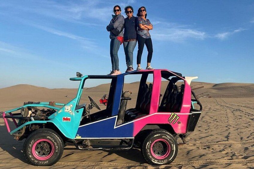 A colorful vehicle with three people standing on top, parked on a sandy terrain with dunes in the background. 3