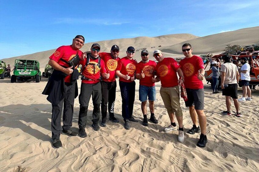 A group of people in red shirts standing on sand dunes with vehicles in the background. 2