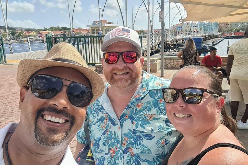 Trois personnes prennent un selfie sur une promenade en bord de mer, avec un port de plaisance et des bâtiments en arrière-plan. 4