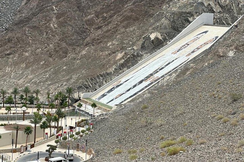 A large dam with a sloped structure, a paved area with palm trees, and rocky terrain. 3