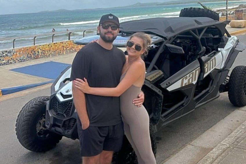 A couple standing next to a black off-road vehicle with 'Can Am' branding, a rocky shoreline, and the ocean in the background. 2